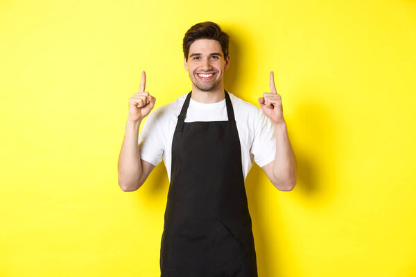 Handsome barista showing advertisement, coffee shop promo offer, pointing fingers up, standing against yellow background