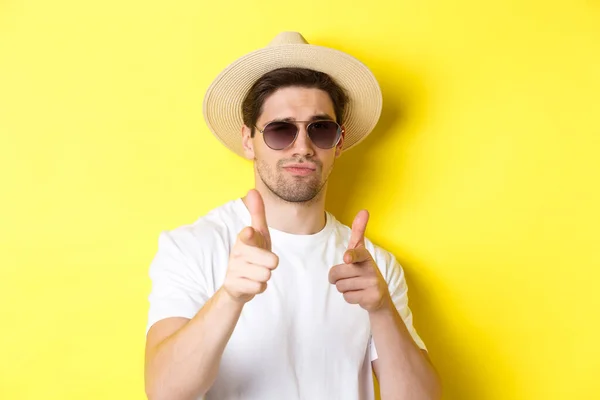 Concepto de turismo y vacaciones. Primer plano de chico fresco en sombrero de verano y gafas de sol apuntando con las armas de dedo a la cámara, de pie sobre el fondo amarillo — Foto de Stock