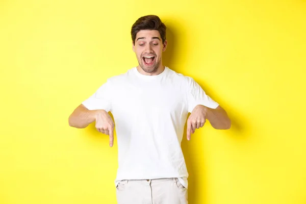 Imagem de cara bonito divertido em t-shirt branca, olhando e apontando os dedos para baixo, de pé sobre fundo amarelo — Fotografia de Stock
