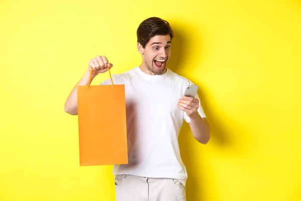 割引、オンラインバンキング、キャッシュバックの概念。黄色の背景を背景に立ちショッピングバッグを見せ、モバイル画面で幸せそうに見える驚きの男 — ストック写真