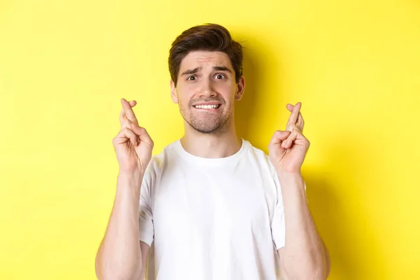 Close-up van nerveuze man maken wens, vingers gekruist en bijtende lip bezorgd, staande over gele achtergrond Stockfoto