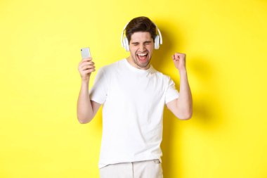 Mutlu adam dans ediyor ve kulaklıkla müzik dinliyor, cep telefonu tutuyor, sarı arka planda duruyor.
