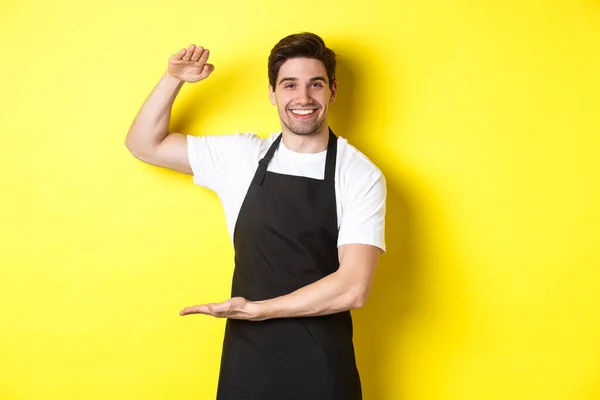 Sorrindo bonito barista mostrando algo longo ou grande, de pé sobre fundo amarelo — Fotografia de Stock