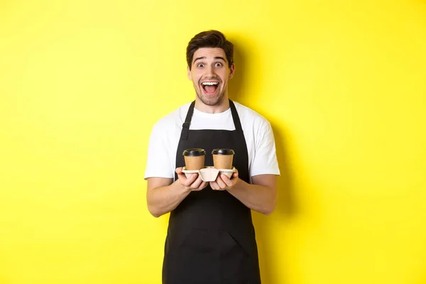 Barista jantan yang gembira memegang dua cangkir kopi lepas landas, bekerja di kafe, berdiri di atas latar belakang kuning — Stok Foto