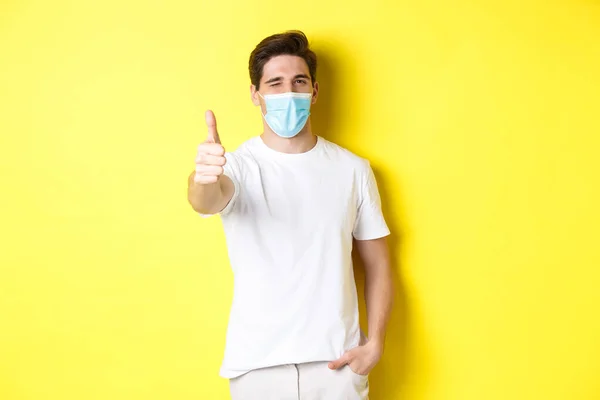 Concept de coronavirus, pandémie et distanciation sociale. Jeune homme confiant dans un masque médical montrant les pouces levés et cligner des yeux, fond jaune — Photo