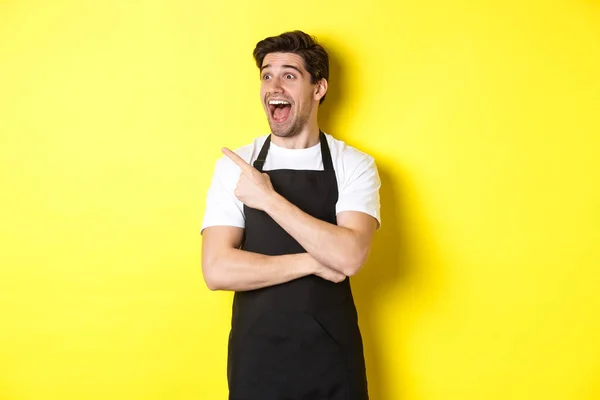 Camarero en delantal negro echa un vistazo a la oferta promocional, señalando el dedo y mirando a la izquierda en su logotipo, de pie sobre el fondo amarillo — Foto de Stock