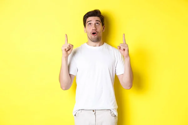 Imponerad kille checka ut promo erbjudande, titta och peka fingrar upp på reklam, står över gul bakgrund — Stockfoto