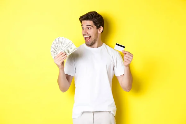 Chico alegre mirando el dinero, la celebración de la tarjeta de crédito, concepto de crédito bancario y préstamos, de pie sobre fondo amarillo — Foto de Stock