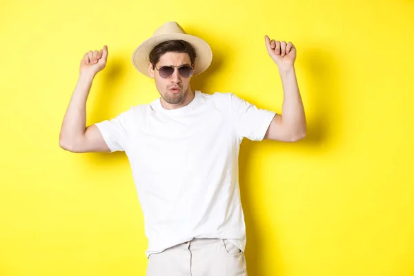 Tourisme, voyage et vacances concept. Touriste homme appréciant les vacances, dansant dans un chapeau de paille et des lunettes de soleil, posant sur fond jaune — Photo