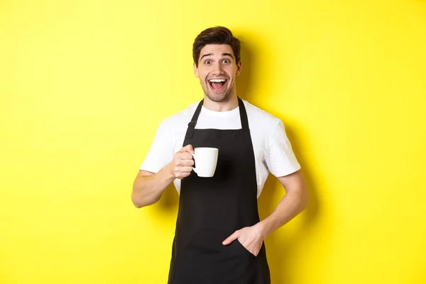 Tersenyum barista muda di celemek hitam memegang cangkir kopi, berdiri di atas latar belakang kuning — Stok Foto