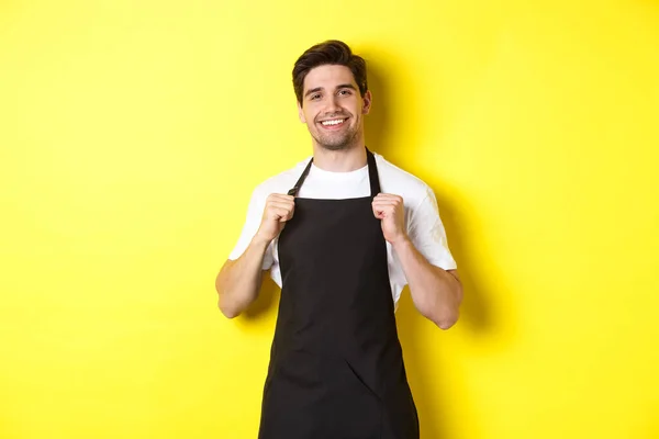 Barista confiante em avental preto em pé contra fundo amarelo. Garçom sorrindo e parecendo feliz — Fotografia de Stock