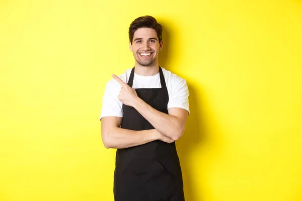 Happy barista menunjuk jari kiri dan tersenyum, mengenakan seragam celemek hitam, berdiri melawan latar belakang kuning — Stok Foto