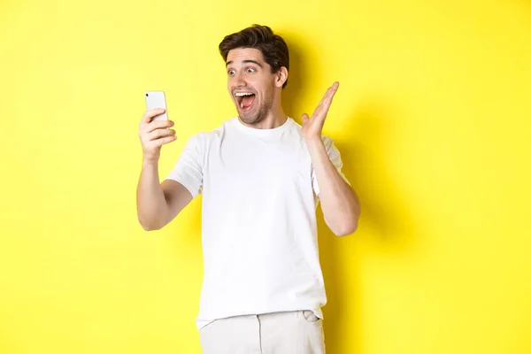 Surpris et heureux homme regardant l'écran de téléphone mobile, la lecture de nouvelles fantastiques, debout sur fond jaune — Photo