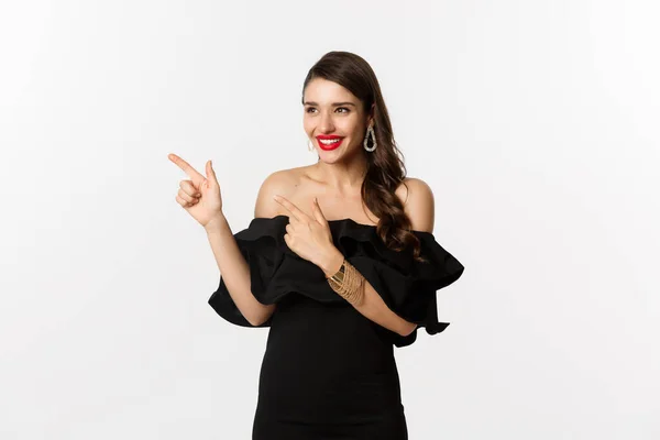 Moda e beleza. Mulher atraente em jóias, maquiagem e vestido preto, rindo e apontando os dedos deixados em oferta promocional, fundo branco — Fotografia de Stock