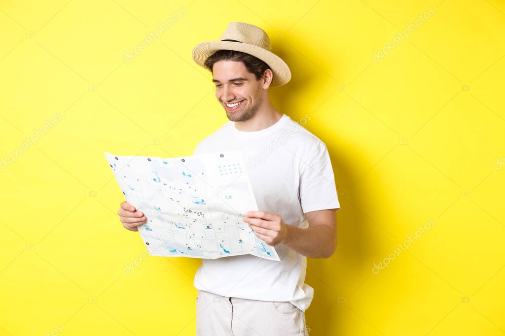 Travelling, vacation and tourism concept. Smiling happy tourist looking at map with sightseeings, standing against yellow background