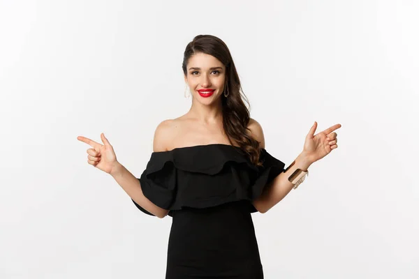 Moda y belleza. Atractiva mujer en joyería, maquillaje y vestido negro, riendo y señalando los dedos hacia los lados ofrecen espacios de copia, fondo blanco — Foto de Stock