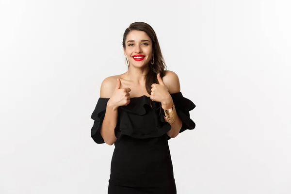 Μόδα και ομορφιά. Πανέμορφη γυναίκα με μαύρο φόρεμα, κόκκινο κραγιόν, δείχνει τους αντίχειρες επάνω στην έγκριση, συνιστώντας προϊόν, στέκεται πάνω από το λευκό φόντο — Φωτογραφία Αρχείου