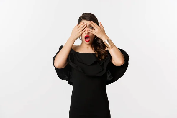 Мода и красота. Изображение шокированной гламурной женщины в черном платье, закрывающей глаза, но выглядывающей сквозь пальцы испуганной, стоящей на белом фоне — стоковое фото