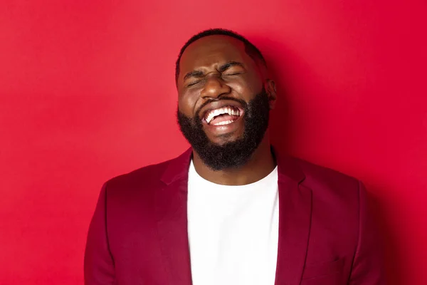 Primer plano del hombre negro feliz y despreocupado divirtiéndose, riendo y sonriendo, de pie en blazer sobre fondo rojo — Foto de Stock