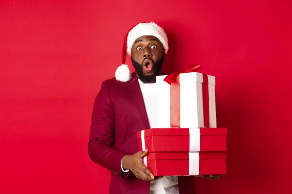 Navidad, Año Nuevo y concepto de compras. Alegre hombre negro secreto santa celebración regalos de Navidad y sonriendo emocionado, traer regalos, de pie sobre fondo rojo — Foto de Stock