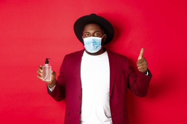 Covid-19, karantina ve tatil konsepti. Tıbbi maskeli, el dezenfektanı ve baş parmağını kaldırmış, antiseptik kullanmayı öneren Afrikalı genç bir Amerikalı.
