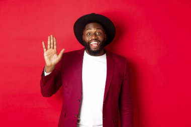 Dost canlısı Afrikalı Amerikalı el sallıyor, merhaba diyor ve gülümsüyor, seni selamlıyor, kırmızı arka planda duruyor.