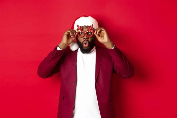 Funny Black man celebrating New Year, wearing party glasses and santa hat, having fun over red background