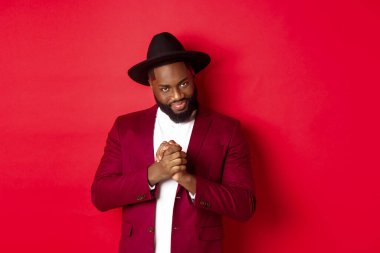 Kurnaz siyah adamın bir fikri var, gülümsemesi sinsi ve ellerini ovuşturuyor, kırmızı arka plana karşı duruyor ve entrika çeviriyor.