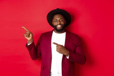 Kış tatili ve alışveriş konsepti. Mutlu siyah adam solu işaret ediyor ve gülümsüyor, kırmızı arka planda yeni yıl tanıtım teklifini gösteriyor.