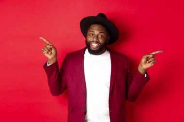 Kış tatili ve alışveriş konsepti. Neşeli siyahi adam gülümsüyor ve iki reklam gösteriyor, fotokopi alanlarını işaret ediyor, kırmızı arka plan