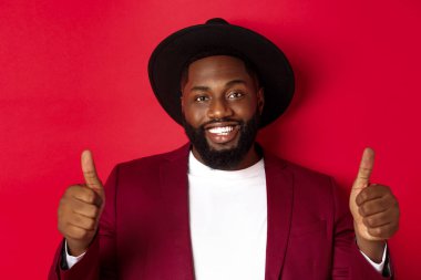 Yakışıklı, sakallı siyahi adamın yakın plan fotoğrafı. Başparmak kaldırıyor, gülümsüyor, memnun, iyi seçimi övüyor, kırmızı arka planda duruyor.