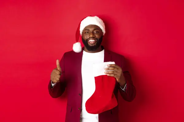 Guapo barbudo chico recibir regalo de Navidad en calcetín, mostrando el pulgar hacia arriba y sonriendo, de pie sobre fondo rojo — Foto de Stock