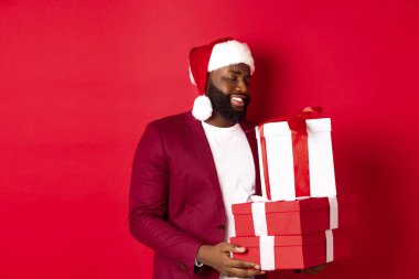 Noel, yeni yıl ve alışveriş konsepti. Mutlu Afrikalı Jamaikalı adam Noel hediyelerine dokunmuş ve minnettar görünüyor, hediyeler alıyor, Noel Baba şapkası takıyor, kırmızı arka plan