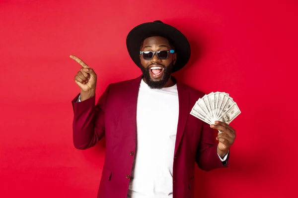 Handsome and stylish Black man pointing fingers left while showing money, holding dollars and demonstrating logo, standing over red background