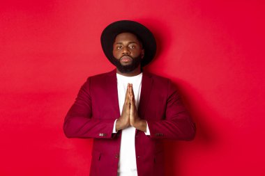 Ciddi Afrikalı Amerikalı adam saygı gösteriyor, namaste 'de el ele tutuşuyor ya da teşekkür ediyor. Kırmızı arka planda duruyor.