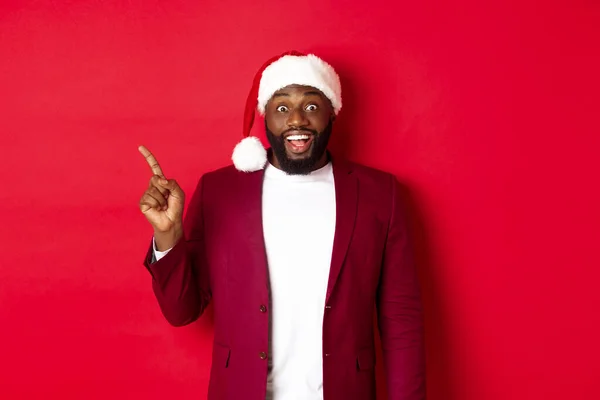 Concepto de Navidad, fiesta y vacaciones. Emocionado y divertido hombre negro apuntando con el dedo hacia la izquierda y sonriendo, mostrando el logotipo, de pie sobre el fondo rojo — Foto de Stock