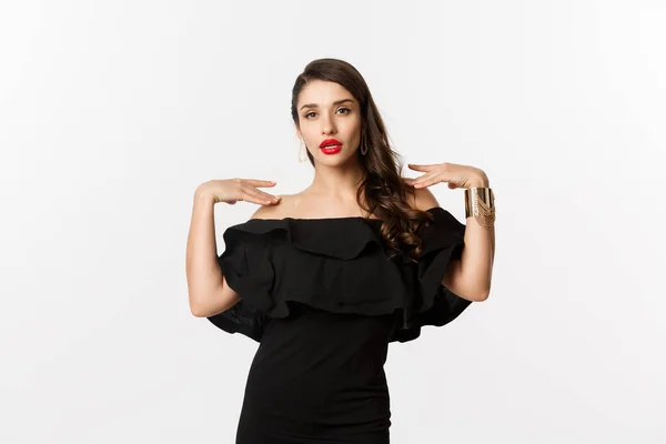Zelfverzekerde en stijlvolle vrouw in zwarte elegante jurk, op zoek sassy op camera, staande over witte achtergrond — Stockfoto