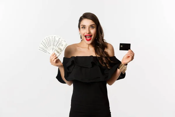 Mode en shopping concept. Opgewonden vrouw in zwarte jurk, tonen credit card en dollars, glimlachen en staren naar de camera, witte achtergrond — Stockfoto