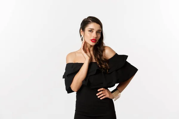 Sensual young woman in black dress, showing her earrings and looking sexy at camera, standing over white background — Stock Photo, Image