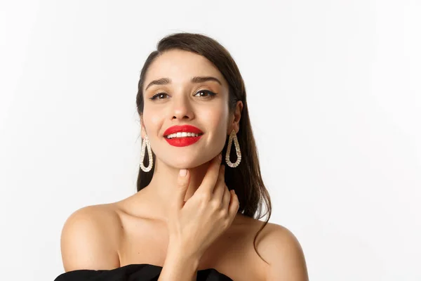 Moda e concetto di bellezza. Primo piano di splendida donna bruna con labbra rosse, viso toccante e sorridente sicuro di sé, in piedi su sfondo bianco — Foto Stock