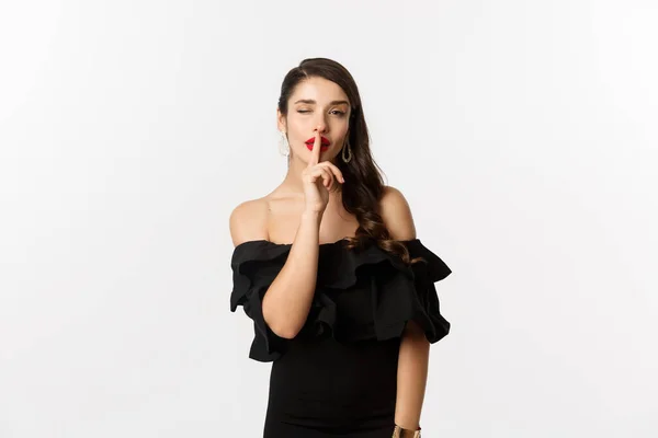 Μόδα και ομορφιά. Coquettish νεαρή γυναίκα σε μαύρο φόρεμα, κόκκινα χείλη, κλείνει το μάτι στην κάμερα και κάνει σήμα σιωπής, στέκεται πάνω από το λευκό φόντο — Φωτογραφία Αρχείου