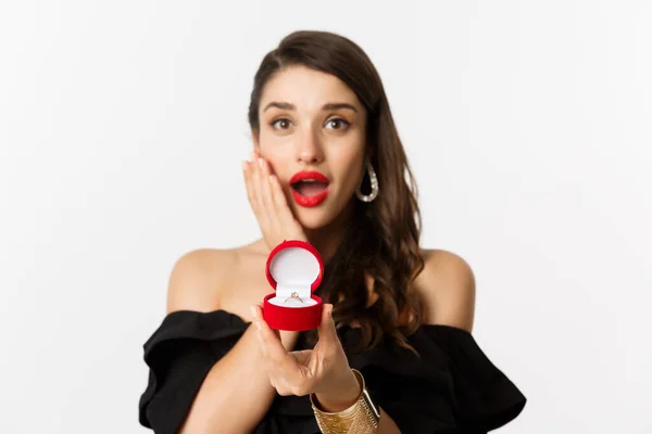 Hermosa mujer en vestido negro haciendo propuesta, mostrando anillo de compromiso y mirando emocionado, de pie sobre fondo blanco —  Fotos de Stock