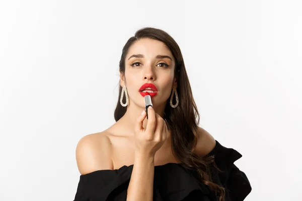 Primo piano di bella donna che applica rossetto rosso sulle labbra, guardando la fotocamera come specchio, in piedi in abito nero su sfondo bianco — Foto Stock