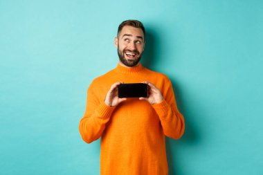 İnternetten alışveriş. Genç adam düşünüyor ve akıllı telefon ekranını gösteriyor, sol üst köşede rüya gibi görünüyor, açık mavi arka planda duruyor.