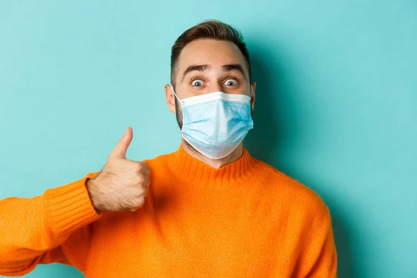 Covid-19, social distancing and quarantine concept. Close-up of cheerful man in medical mask showing thumb-up, light blue background — Stock Photo, Image