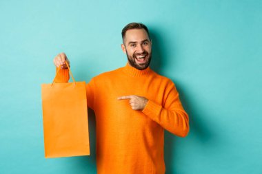 Tatmin olmuş erkek müşteri turuncu alışveriş torbasını işaret ediyor, mağazayı tavsiye ediyor, gülümsüyor, turkuaz arka planda duruyor.