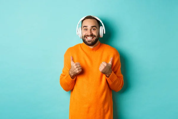 Verbazingwekkende volwassen man luisteren muziek in hoofdtelefoon, kijken naar de camera onder de indruk en het tonen van duimen-up in goedkeuring, aanbevelen, staande over turquoise achtergrond — Stockfoto