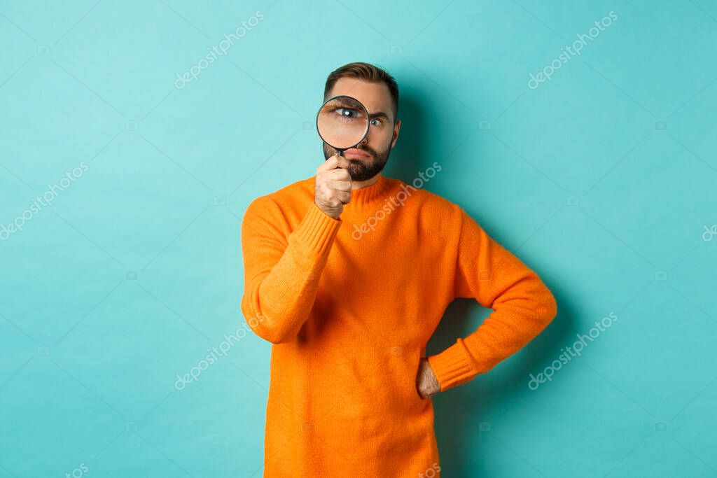 Funny man looking serious through magnifying glass, inspecting something, standing in orange sweater against turquoise background.