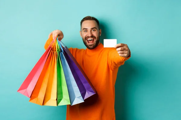 幸せな男を示すクレジットカードとショッピングバッグ、ターコイズの背景に立って — ストック写真