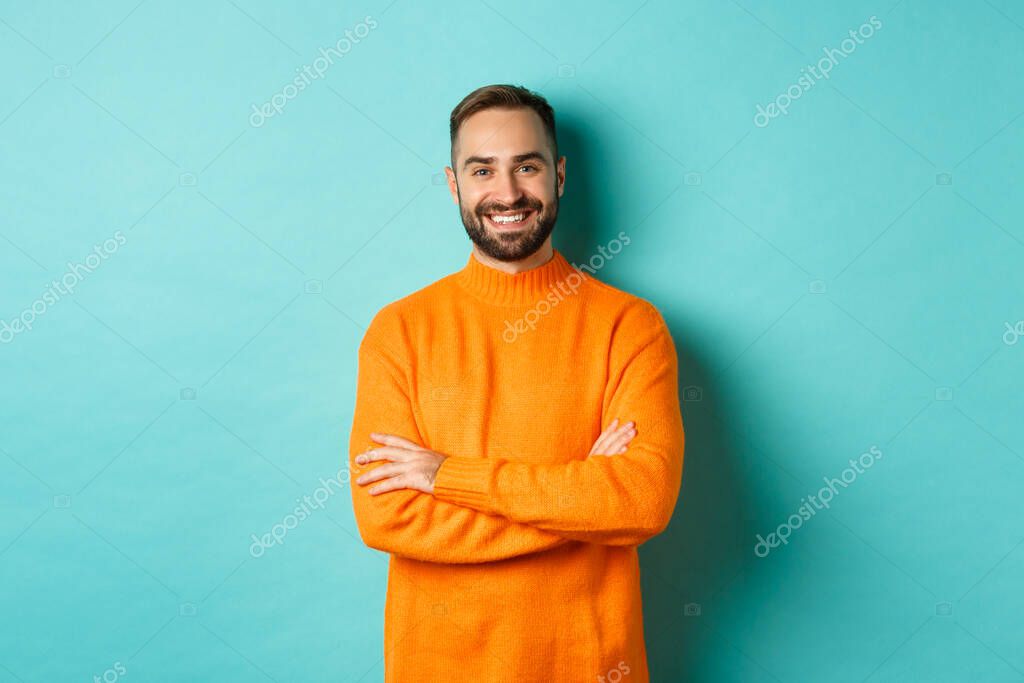 Handsome bearded male model in orange sweater, looking confident and smiling, cross arms on chest, standing over turquoise background.